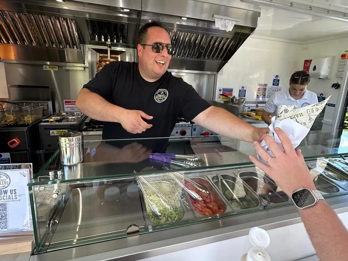 A Somerset Sizzlers chef handing a freshly made wrap across the counter, with the gyros spit turning behind him
