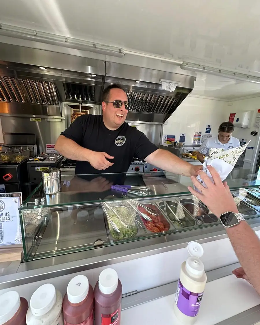 A Somerset Sizzlers chef in sunglasses handing a wrap across the counter, with the gyros spit turning behind him