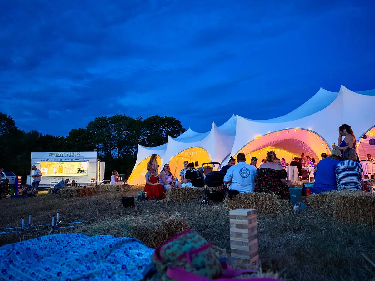 The Somerset Sizzlers trailer glowing at dusk beside a lit stretch marquee, with guests relaxing on hay bales